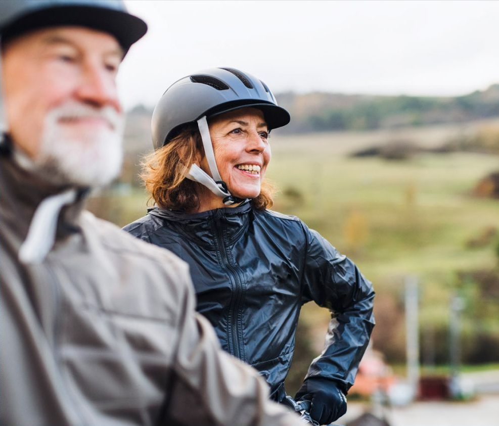 Close-up of a smiling woman in a black helmet and jacket outdoors with another blurred figure wearing a helmet in the foreground in rathdrum id