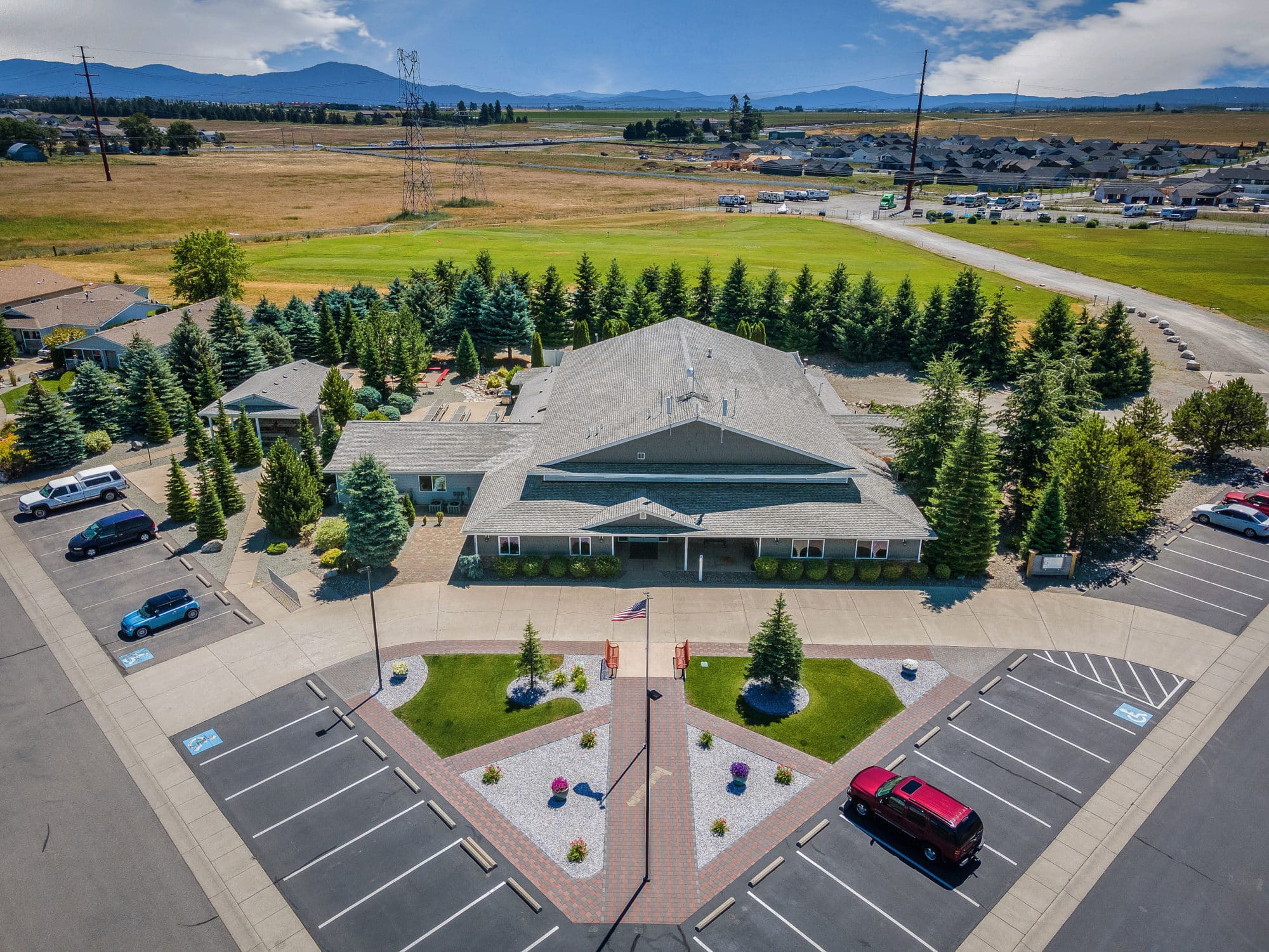 Elevated view of a large community building with a parking lot, surrounded by landscaped greenery and open fields with mountains in the distance in rathdrum id