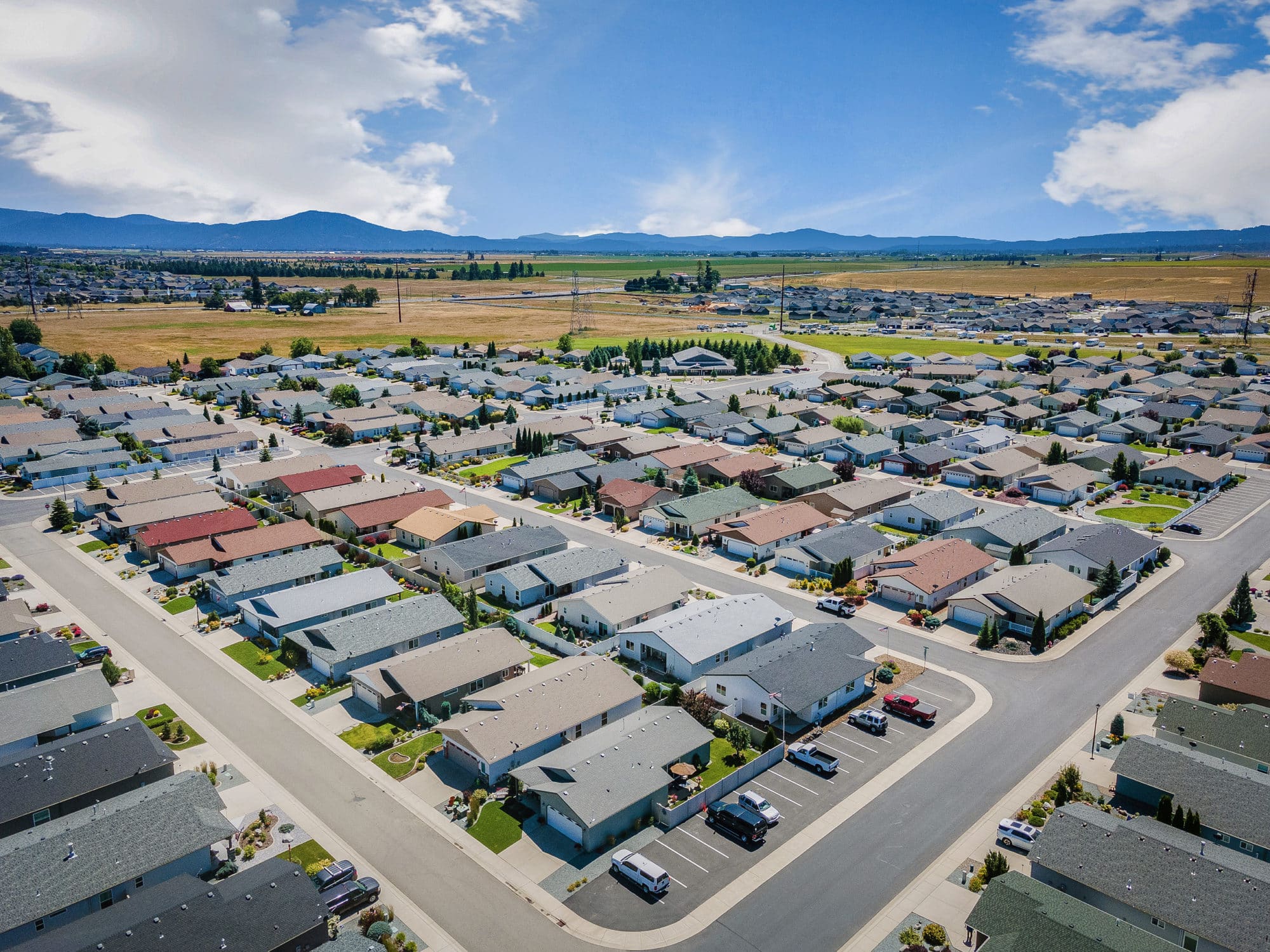 Aerial view of a residential community with neatly arranged homes, landscaped yards, and surrounding open fields and mountains under a bright sky in rathdrum id