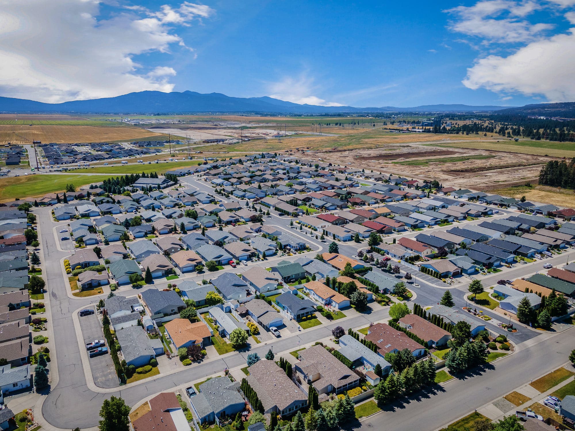 Wide aerial view of a residential community surrounded by open fields, mountains, and scattered trees in rathdrum id