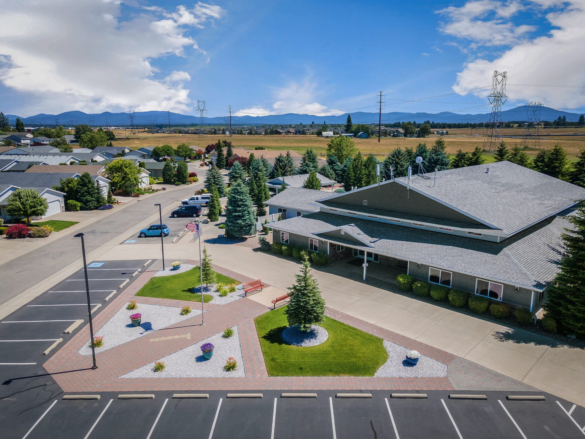 Aerial view of a building with a gray roof, a landscaped entrance with benches and a flagpole, surrounded by a parking lot and suburban area in rathdrum id